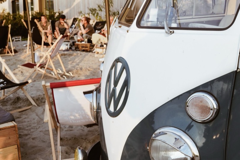 Nahaufnahme VW-Emblem am Bulli, im Hintergrund Gäste in Strandstühlen