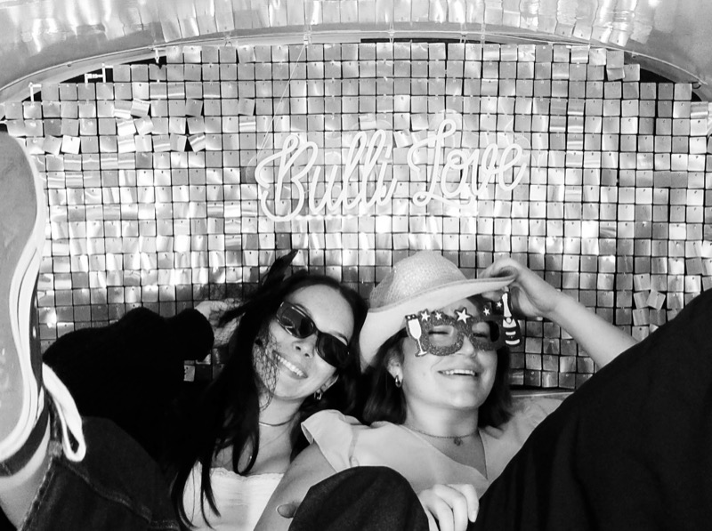 Zwei Frauen mit Sonnenbrille und Hut vor der Bulli Love Pailletten-Fotowand